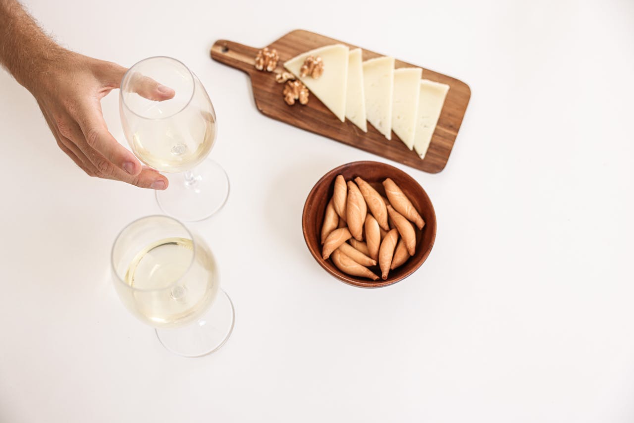 Top view of cheese slices, breadsticks, and wine glasses on a white surface for an elegant dining setup.